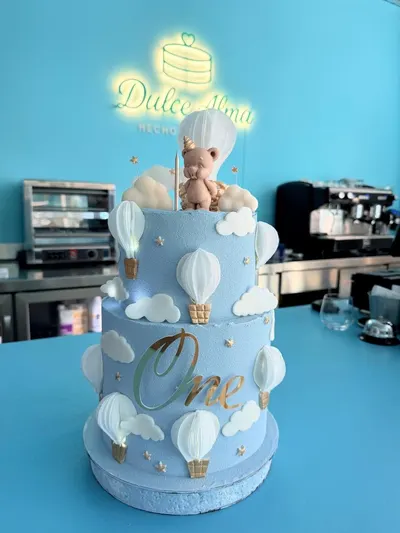 Tarta de dos pisos azul cielo con globos aerostáticos, nubes y un osito, frente al rótulo de Dulce Alma